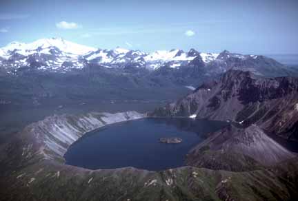 Photograph from air of volcano with lake in crater and snow-covered peak in far background