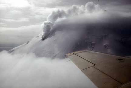 Photograph from air of steam from volcano mixed with clouds; airplane wing in foreground