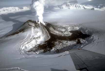 Photograph from air of dark lava flow melting into ice-filled caldera at base of steaming cone
