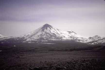 Photograph of partially snow-covered, conical volcano in distance with flat, debris fan in foreground