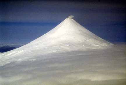 Photograph from air of perfectly conical, snow-covered, steaming volcanic peak and clouds