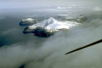 Photograph from air of snow-covered, steep-sided island volcano with ocean, clouds, and plane wing in foreground