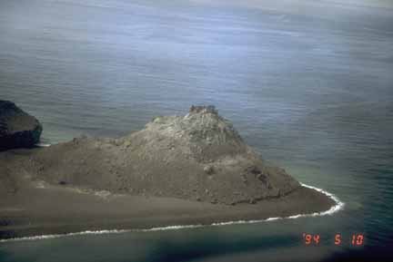 Photograph from air of dark lava dome and beach surrounded by blue sea