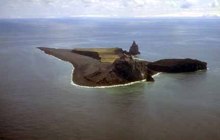 Photograph from air of dark, jagged volcanic island surrounded by calm ocean, clouds on horizon