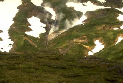 Photograph of grassy slopes, snow patches, and steaming areas near yellow-stained rocks
