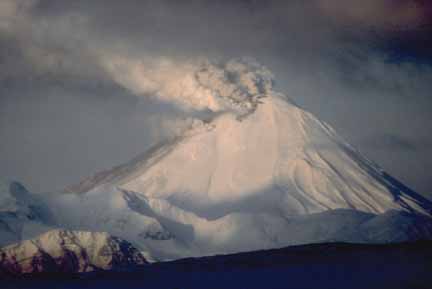 Photograph of gray ash and steam coming from top of snow-covered volcanic cone, steep ridges in middle ground
