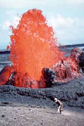 photo 083.  Photo of red-hot lava fountaining in middleground with geologist wearing heat-shielding clothing and full-face mask collecting samples from quite nearby