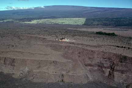 photo 099.  Low-elevation oblique aerial photo of edge of crater in middleground with buildings perched on the edge